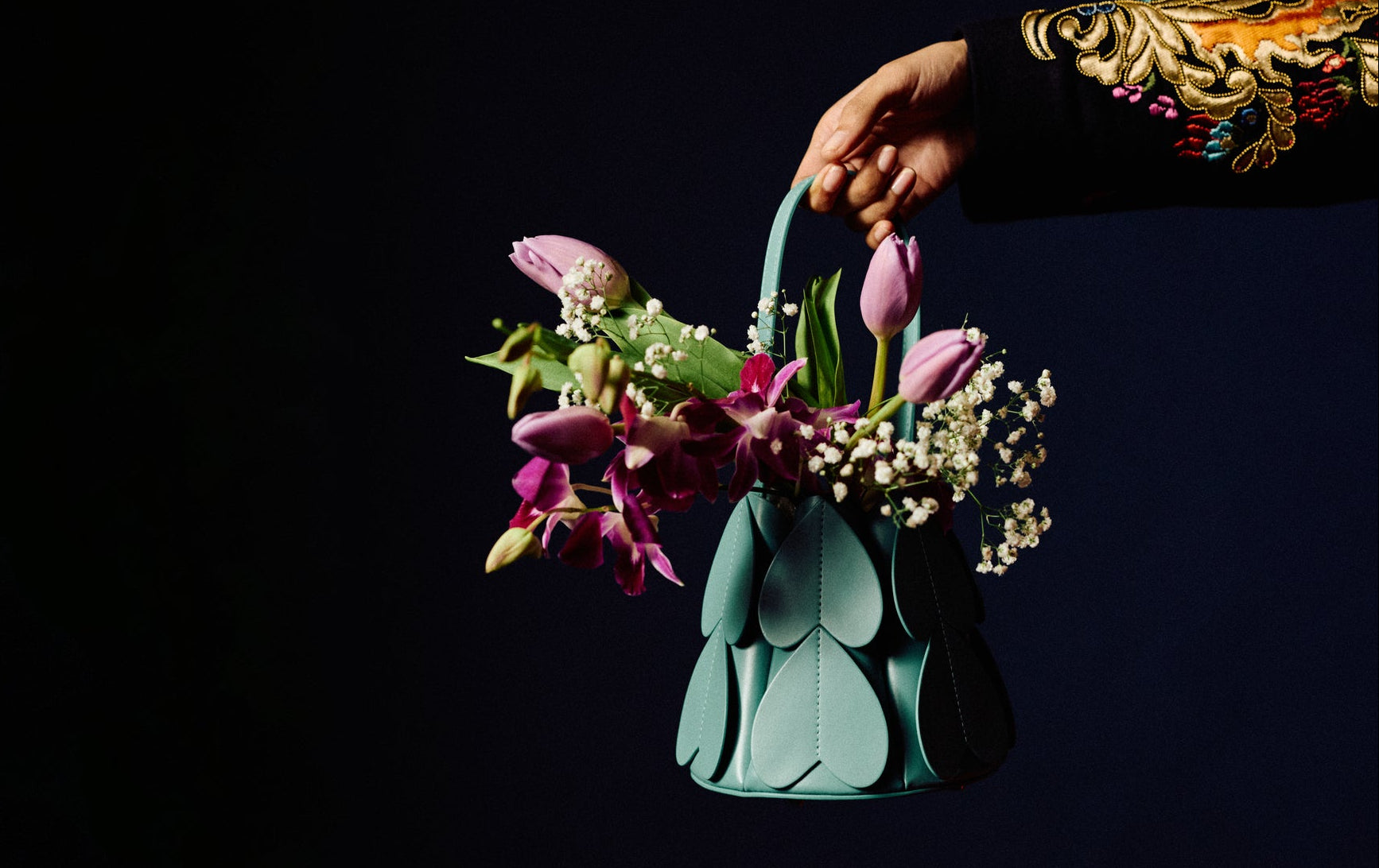 Hand holding a turquoise handbag filled with flowers against a black background